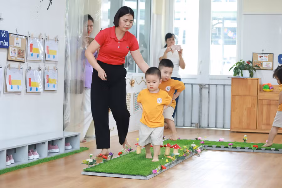 Cô giáo hướng dẫn trẻ lớp Nhà trẻ hoạt động phát triển vận động đi bước qua chướng ngại vật.
