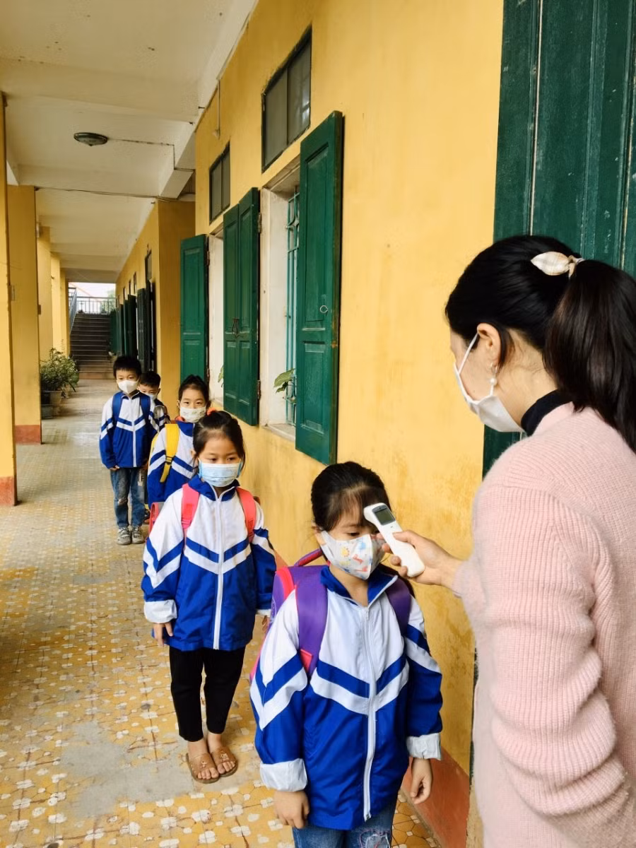 Cô giáo đo thân nhiệt cho học sinh tại của lớp học.