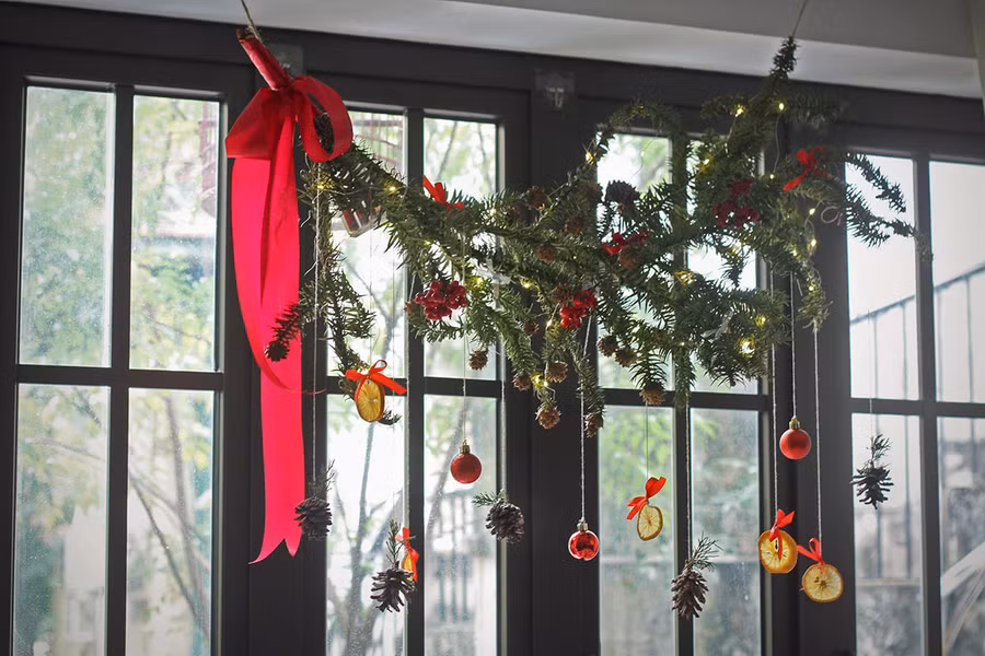 1 bó cành thông thật làm được 2 chiếc vòng, còn thừa treo ngang lên thanh rèm cửa sổ rồi treo đồ trang trí, đèn led chạy pin và ruy-băng lên trên.