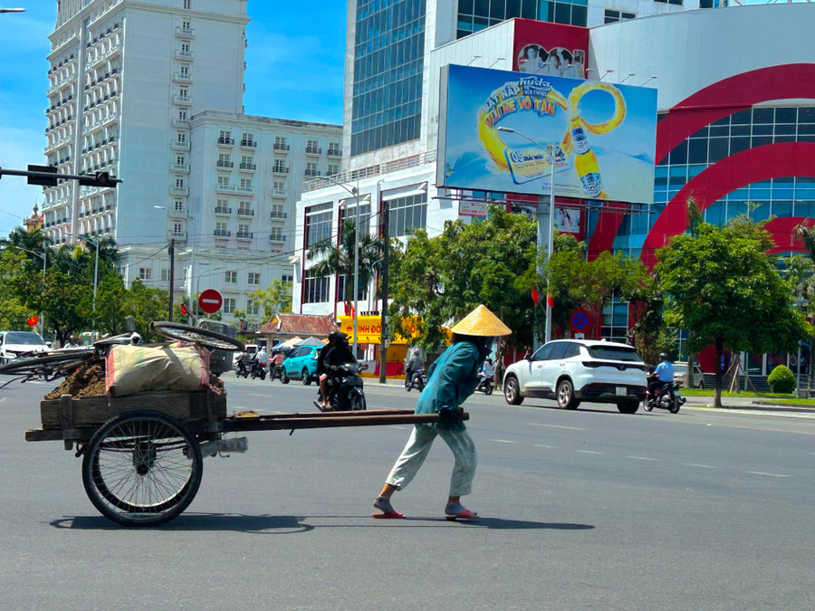 Một phụ nữ trung niên cúi người kéo xe đất nặng trĩu trên mặt đường nóng rát, mồ hôi đầm đìa sau lưng áo. anh-6.jpg