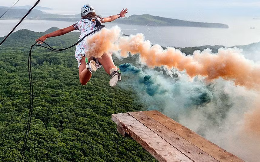 Elena Kazachenko jumps from a wooden beam with a smoke flare attached to her ankle. The adrenaline junkie can be seen leaping from a makeshift wooden beam precariously placed over the edge of an an electricity pylon. However, instead of plummeting 300 feet to the ground below, she is suddenly snatched to safety by a self-made zip line. Elena, from Russia, even leaves behind a stream of coloured smoke from a signal flare attached to her ankle.