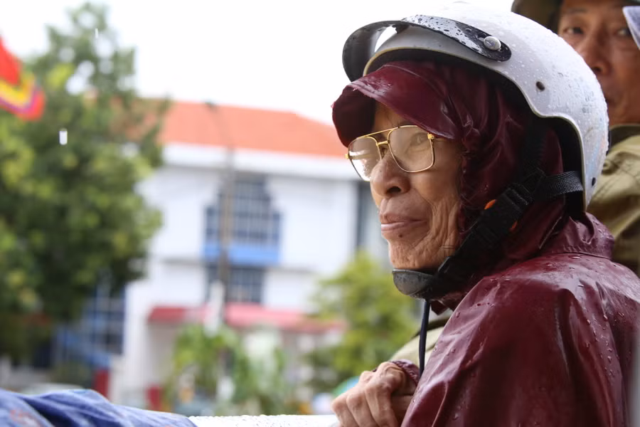 Cụ bà đội mưa theo dõi từng thuyền đua trên sông.