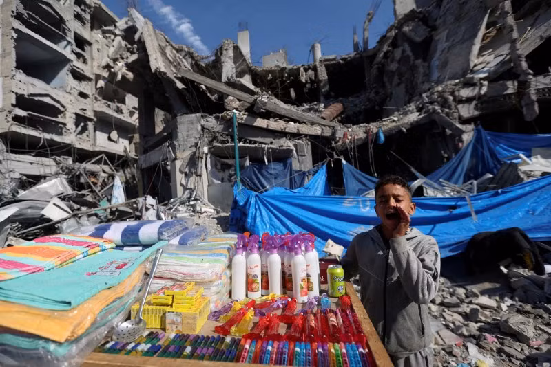 Một người bán hàng Palestine chào bán sản phẩm của mình tại một khu chợ trời gần đống đổ nát trong các cuộc tấn công của Israel. Chùm ảnh gần 40.000 tòa nhà bị phá hủy ở Gaza