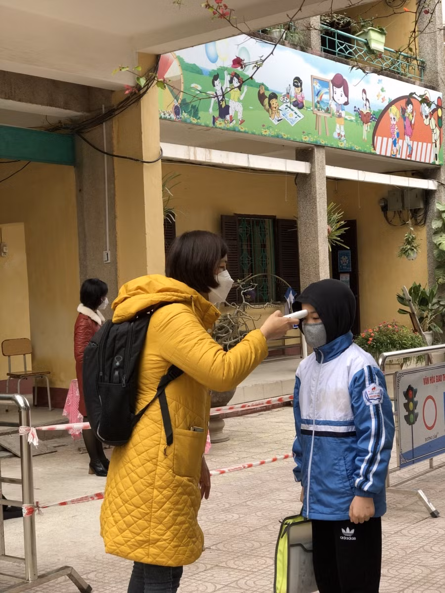 Giáo viên đứng hướng dẫn và hỗ trợ đo thân nhiệt cho học sinh.