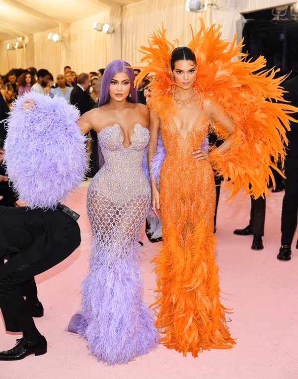 Chị em nhà Jenner chơi màu nổi. Kylie (trái) diện đầm lưới tím của Versace, còn Kendall chọn đầm xẻ ngực kèm áo choàng lông vũ màu cam neon.