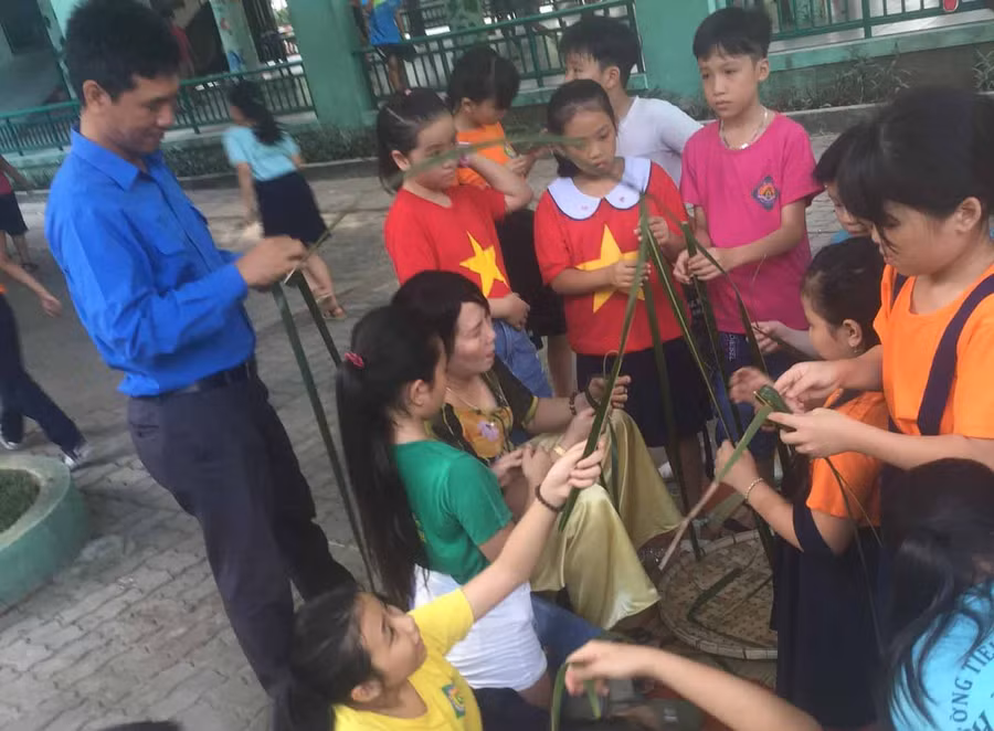 Học sinh chăm chú xem cô giáo hướng dẫn cách thắt chong chóng, châu chấu từ lá dừa. Ảnh: NVCC