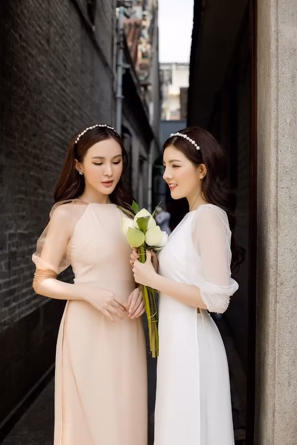 Cặp chị em thân thiết Kelly và Lily Luta nền nã trong tà áo dài cổ tròn màu be - trắng với phần tay voan bồng.