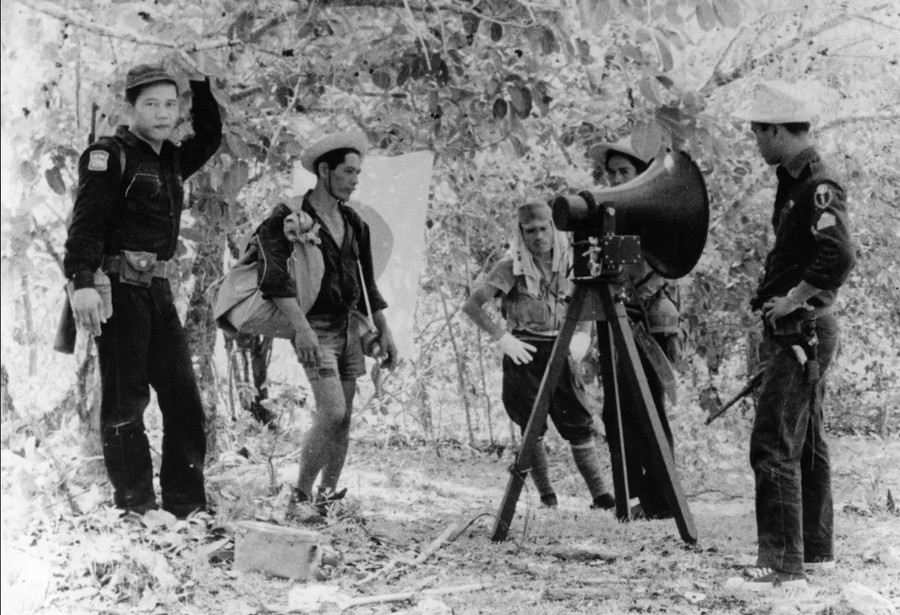 Binh lính Nhật Bản và cảnh sát Philippines đứng cạnh loa phóng thanh cung cấp thông tin về gia đình các binh sĩ Nhật Bản, Lubang, Philippines, năm 1959.