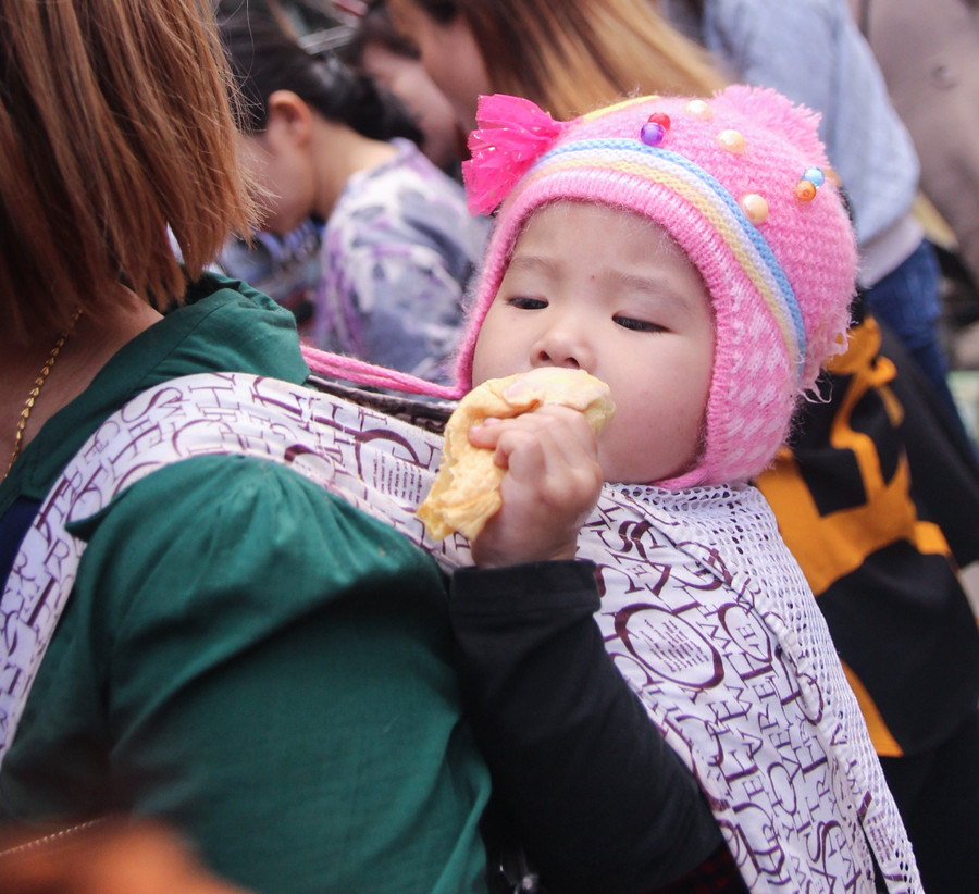 Em bé được mẹ địu sau lưng đi chợ, mải mê với chiếc bánh của mình