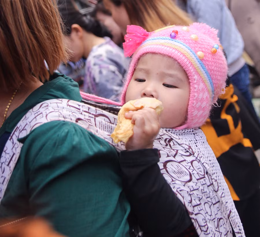 Em bé được mẹ địu sau lưng đi chợ, mải mê với chiếc bánh của mình