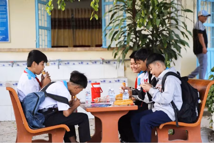 Các em học sinh vui vẻ ăn bánh mì trong sân trường và tranh thủ đọc tờ rơi tuyên truyền. Ảnh: CACC.