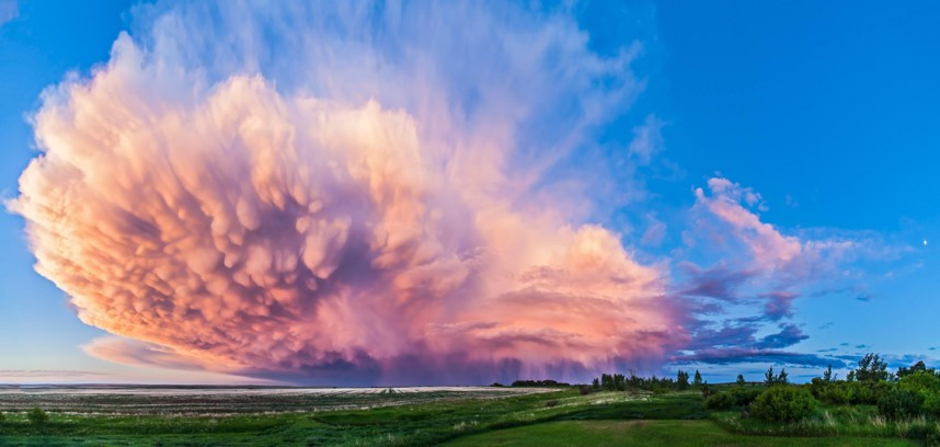 A huge storm cloud glowing at sunset in Canada. The storm itself is visible at the far end of the cloud in the distance, with bulbous mammatus cloud formations in the foreground.