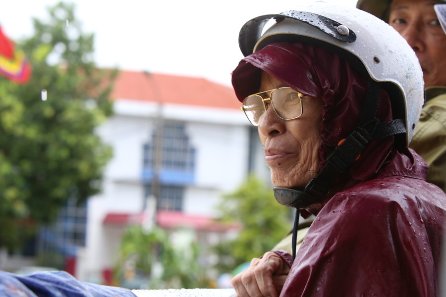 Cụ bà đội mưa theo dõi từng thuyền đua trên sông.