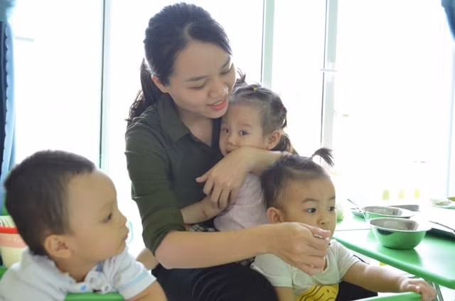 Giờ ăn của nhóm trẻ 6 - 12 tháng tuổi tại Trường Mầm non Hoàng Cúc (Q. Sơn Trà, TP Đà Nẵng), GV vừa phải bồng trẻ vỗ về vừa cho các bé khác ăn. 
