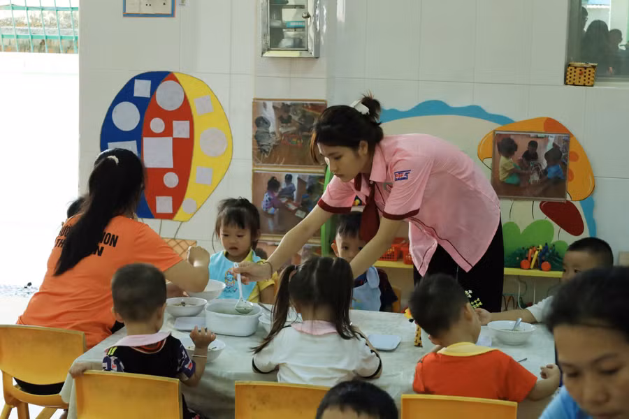 Trần Thị Diễm Quỳnh, sinh viên ngành Giáo dục Mầm non (áo sơ mi hồng) phụ các cô giáo lớp nhà trẻ ở Trường Mầm non Thạnh An cho các bé ăn trưa.